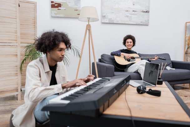 African american woman playing acoustic guitar near blurred boyfriend with synthesizer and laptop at home  - Foto, Imagem