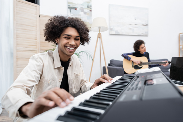 Smiling african american man playing synthesizer near laptop and blurred girlfriend with acoustic guitar at home  - 写真・画像
