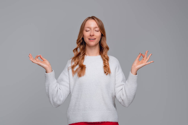 Zen. Portret spokojnej młodej blondynki medytującej, podnoszącej ręce w geście mudry, zamykającej oczy, starającej się zrelaksować po długich godzinach nauki, noszącej biały sweter, stojącej na szarym tle - Zdjęcie, obraz