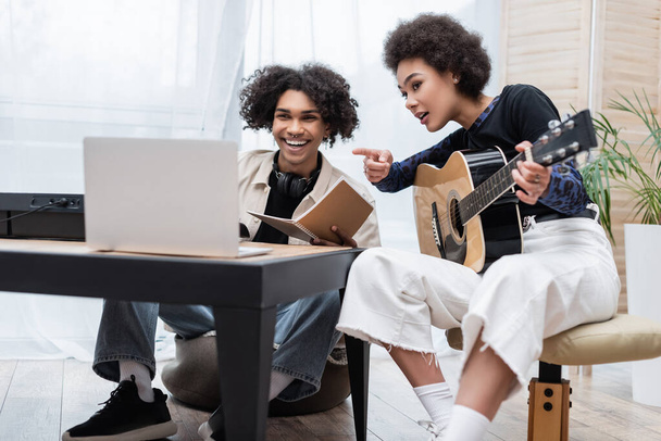 Smiling african american man looking at blurred laptop near synthesizer and girlfriend with acoustic guitar at home  - Foto, Imagen