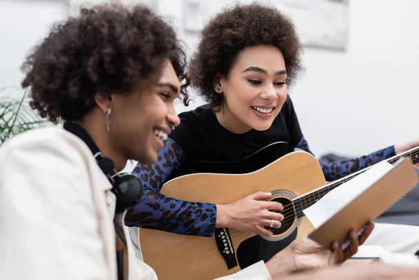 Young african american man holding notebook near girlfriend playing acoustic guitar at home  - Foto, imagen