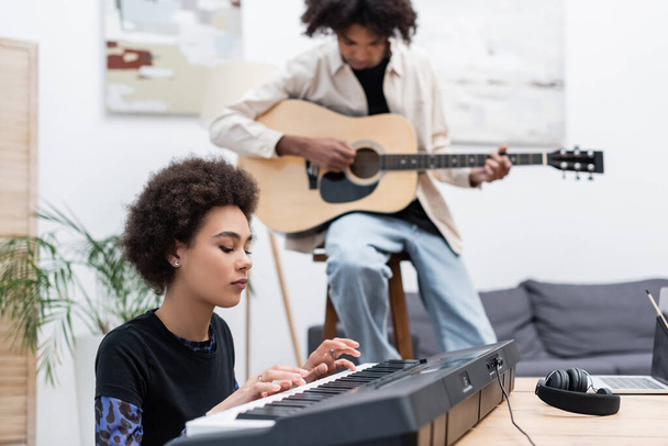 Afroamerykanka gra w syntezatora w pobliżu laptopa i niewyraźny chłopak z gitarą akustyczną w domu  - Zdjęcie, obraz