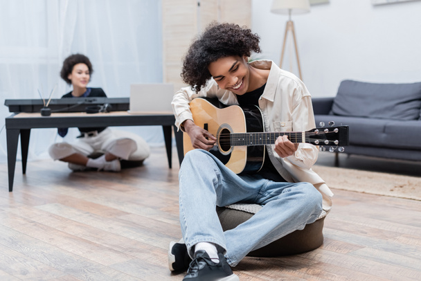 Cheerful african american man playing acoustic guitar near blurred girlfriend at home  - Foto, Imagem