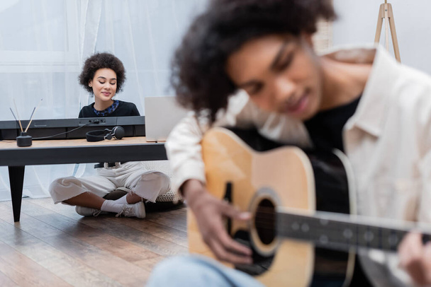 Smiling african american woman sitting near laptop, synthesizer and blurred boyfriend playing acoustic guitar at home  - Foto, imagen