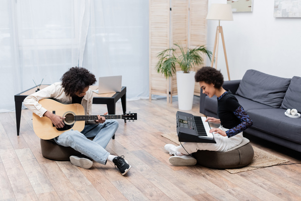 Mosolygó afro-amerikai pár játszik akusztikus gitár és szintetizátor a nappaliban  - Fotó, kép