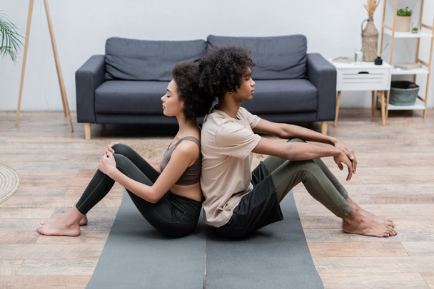 Vista lateral de una joven pareja afroamericana meditando espalda con espalda sobre esterillas de yoga en casa  - Foto, Imagen