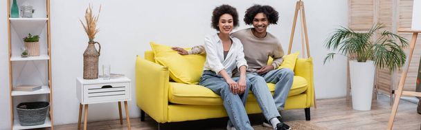 Χαρούμενο ζευγάρι Αφροαμερικανών που κοιτούν την κάμερα στον καναπέ κοντά στο καβαλέτο στο σπίτι, πανό  - Φωτογραφία, εικόνα