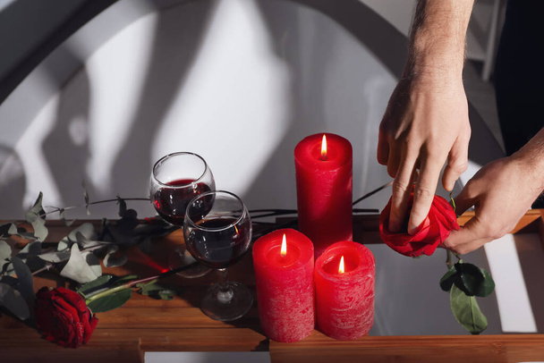 Uomo con rosa rossa vicino vassoio con candele aroma e bicchieri di vino in bagno in data romantica - Foto, immagini