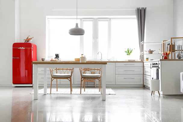 Interior de la cocina moderna y luminosa con nevera roja, mostradores blancos y mesa de comedor - Foto, Imagen