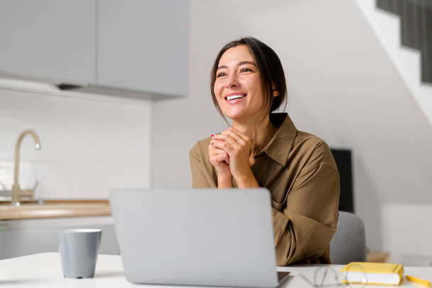 Smiling young cheerful asian woman enjoying morning coffee while working on the laptop, sitting at the desk in the home office. Happy businesswoman - Foto, Imagen
