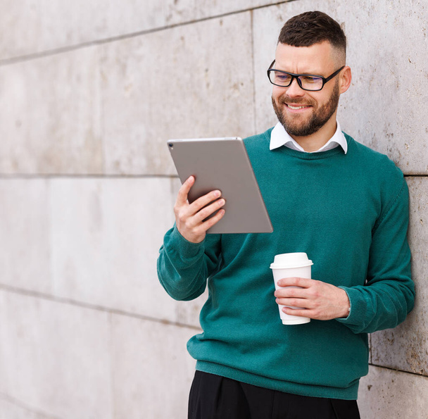 Lachende jonge kantoormedewerker in casual kleding buiten staan en met behulp van digitale tablet - Foto, afbeelding