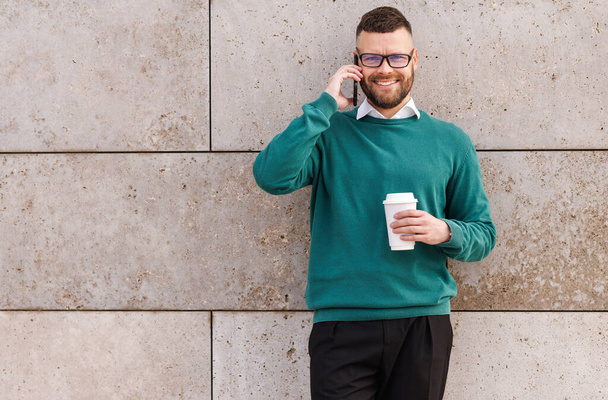 Junger fröhlicher Büroangestellter draußen telefoniert mit Kaffee zum Mitnehmen in der Hand - Foto, Bild