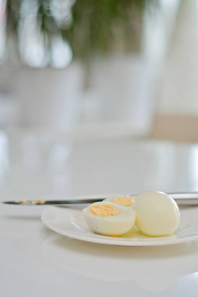 Uova sode su un tavolo di cucina, colazione sana e dieta ricca di proteine. Uova su un piatto con un coltello su un lato, sfondo arioso e bianco con piante verdi e un riflesso luminoso dal tavolo. - Foto, immagini