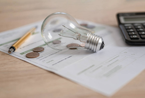 Účet za elektřinu s žárovkou, několika mincemi, kalkulačkou a perem na stole. Koncepce cen elektřiny a daňových plateb. - Fotografie, Obrázek