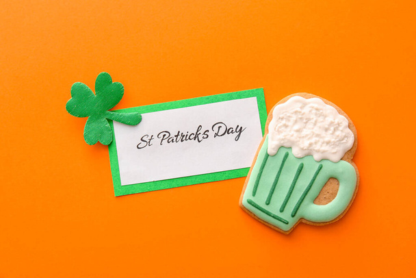 Tasty gingerbread cookie for St. Patrick's Day celebration and card on orange background - Fotó, kép