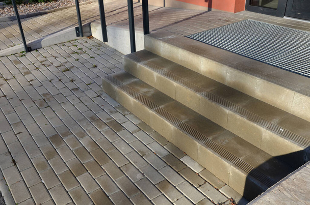 betonowe schody z antypoślizgowymi rowkami. obok schodów jest rampa dla wózków inwalidzkich przy wejściu do mieszkania, dom. metalowa siatka do czyszczenia obuwia we wnęce w płytkach - Zdjęcie, obraz