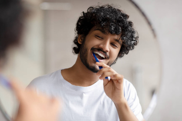 Oral care concept. Young indian man cleaning teeth with toothbrush, smiling to his reflection in mirror - 写真・画像