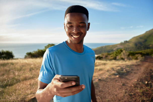 Μαύρος νεαρός ενήλικας χαμογελάει κοιτάζοντας το κινητό του. Στέκεται έξω στο βουνό του. - Φωτογραφία, εικόνα