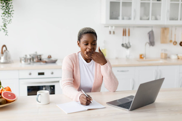 Tired young pretty black female looks at laptop makes notes, yawns at minimalist kitchen interior, free space - Foto, Imagem