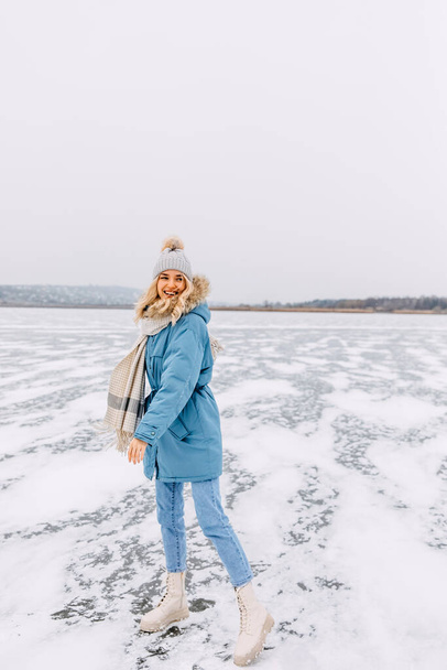 Feliz joven rubia de pie sobre hielo en un lago congelado en un frío día de invierno. - Foto, imagen
