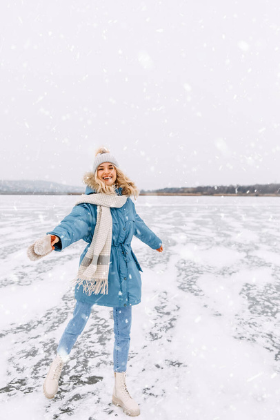 Cheerful young woman dancing on ice on a frozen lake on a cold winter day in snow fall. - Photo, image
