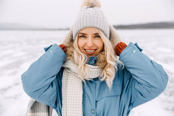Portrait of a cheerful young blonde woman on cold winter day, smiling, adjusting hat. - Photo, Image