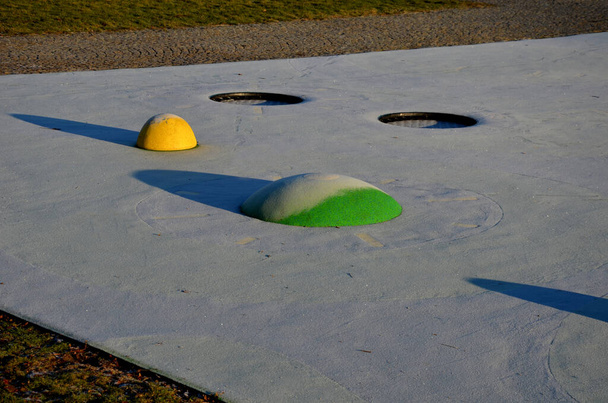 weiße Gummioberfläche eines Spielplatzes mit gelben Schaufeln und farbigen Flecken im Frost menschlicher Fußabdrücke. Erhöhte Linsen wie Bälle, Holzbänke und runde Trampoline im Gelände versenkt. - Foto, Bild