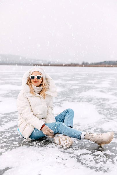 Happy young blonde woman sitting on frozen lake in winter. - Photo, image