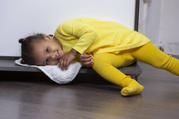 Una niña afroamericana con un vestido amarillo yace en el suelo y sonríe. - Foto, Imagen