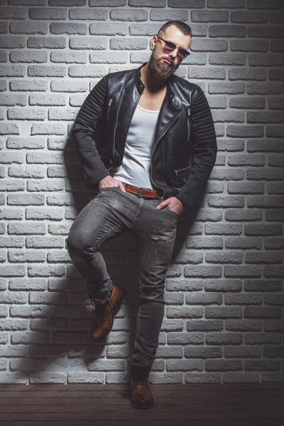 Attractive stylish man with beard in leather jacket and sunglasses looking at camera, standing against brick wall - Photo, Image