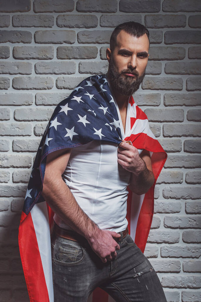 stylischer Mann mit Bart, bedeckt mit amerikanischer Flagge wie ein Superheld, in die Kamera blickend, vor Backsteinmauer stehend - Foto, Bild