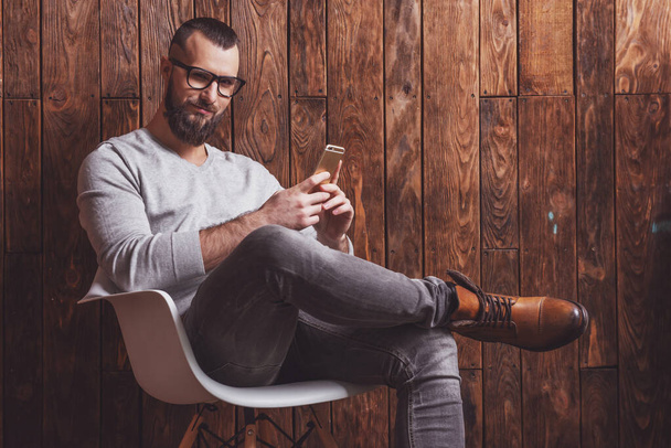 Stylish man with beard in gray sweater and eyeglasses using a mobile phone, sitting on a chair against wooden background - Photo, Image