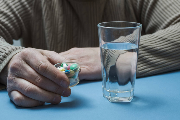 L'homme tient des pilules dans ses mains, il y a un verre d'eau devant lui. - Photo, image