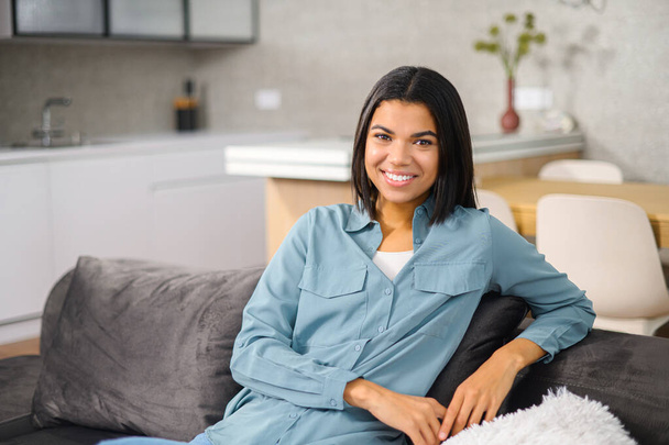 Carefree multiracial woman resting on the comfortable couch at home, while feeling happy. Charming woman looking at camera and smiling while resting - Foto, immagini