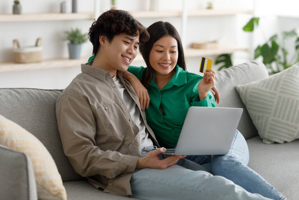 Pozitív ázsiai család csinál online vásárlás hitelkártyával és laptoppal, ül a kanapén és ölelés otthon - Fotó, kép