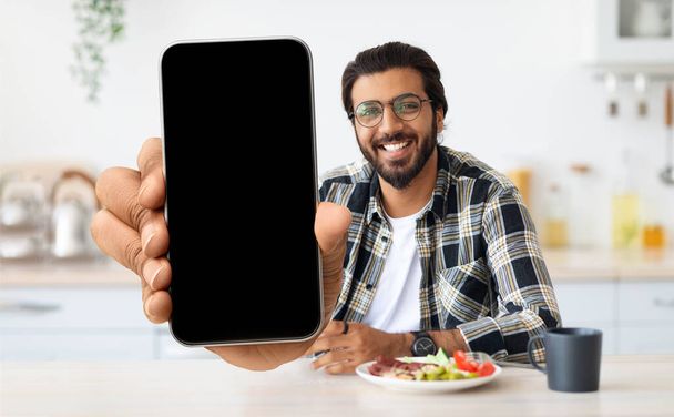 Bonito indiano homem mostrando em branco Smartphone na câmara enquanto sentado na cozinha - Foto, Imagem