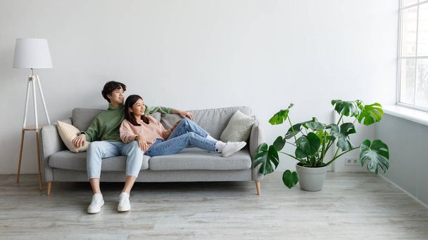 Positivo giovane coppia asiatica rilassante sul divano, coccole e guardando fuori dalla finestra in soggiorno, spazio libero - Foto, immagini