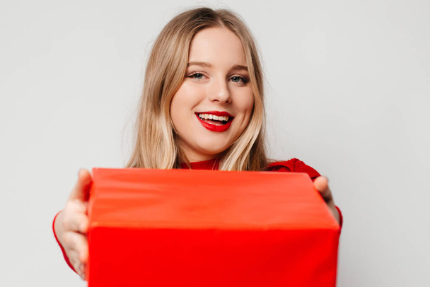 Młoda kobieta w czerwonym swetrze z czerwonymi ustami dająca aparatowi czerwone pudełko prezentów. Kobieta daje dar. - Zdjęcie, obraz
