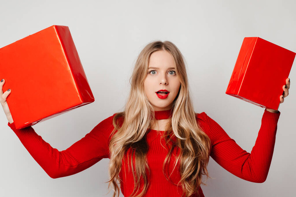 Young blonde woman in red holding gifts in both hands looking at the camera with doubtful expression - Foto, imagen