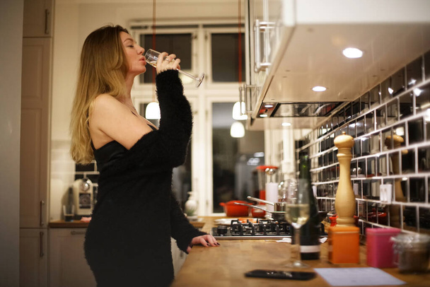 молодая женщина в черном коктейльном платье пьет шампанское на кухне                                                            - Фото, изображение