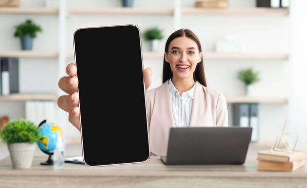 Mobile Attrappe. Lächelnde Dame demonstriert großes leeres Smartphone, während sie im Büro sitzt - Foto, Bild