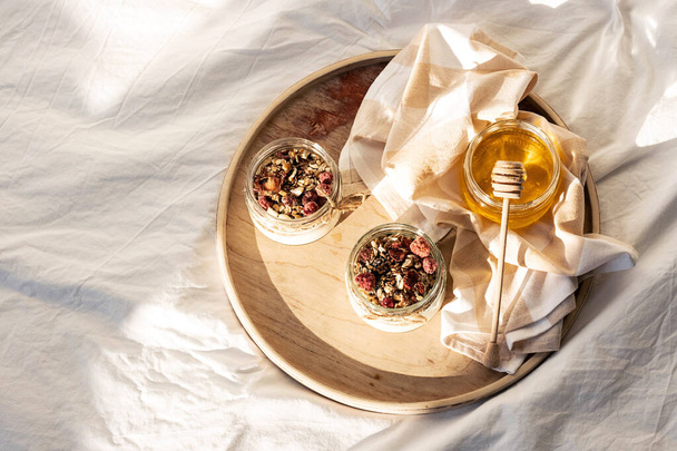 Hausgemachtes Müsliparfait. Schnelles gesundes Frühstück griechischer Joghurt, Honig, Müsli mit getrockneten Beeren und Nüssen im Glas auf Holztablett in weißer Bettwäsche in der Morgensonne.  - Foto, Bild