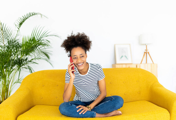 Cheerful millennial female talking on smartphone while sitting on sofa at home - Photo, Image