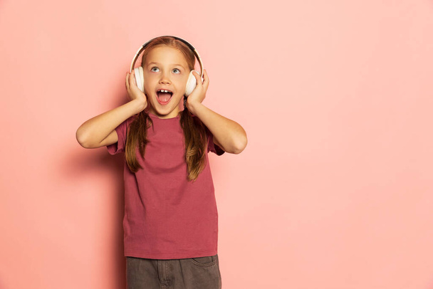 Happy little girl wearing t-shirt listening to music in headphones isolated on pink background. Concept of childhood, emotions, study - 写真・画像