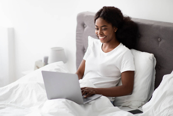 Alegre contratista independiente afroamericana dama usando portátil en la cama - Foto, imagen