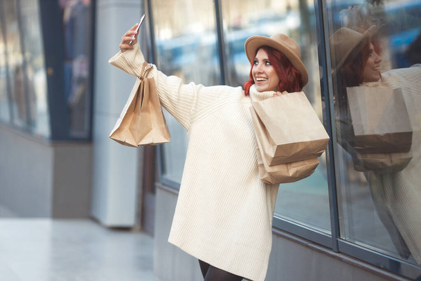 Bin yıllık mutlu moda kadının sokak tarzı portresi. Alışveriş yaptıktan sonra bir sürü el işi çantasıyla cep telefonundan sosyal medya için selfie çekiyor ve alışveriş merkezinin girişinin yanında duruyor.  - Fotoğraf, Görsel