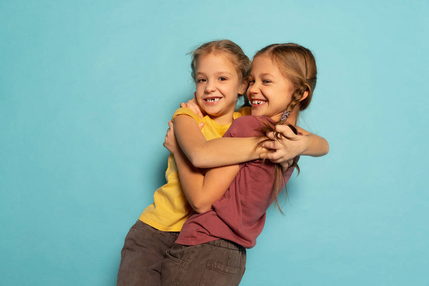 Studio shot of cute little girls, sisters or siblings wearing casual style clothes isolated on blue background. Concept of childhood, emotions - Фото, зображення