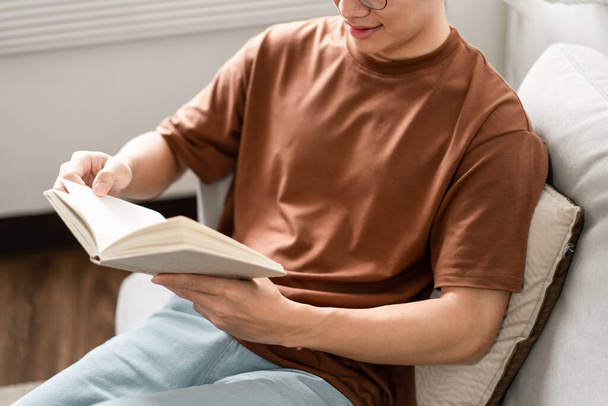 Technologia Koncepcja Samiec w okularach siedzi na szarej sofie, opierając się o nią plecami i czytając książkę. - Zdjęcie, obraz