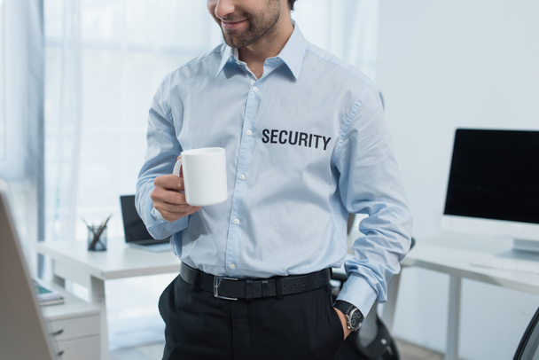 vista ritagliata di sorridente uomo di sicurezza con una tazza di tè in piedi vicino a monitor sfocati in ufficio - Foto, immagini