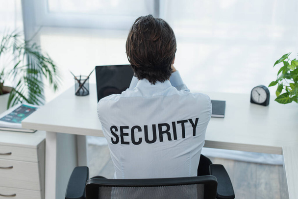 Вид сзади охранника в рубашке с наручниками, сидящего рядом с ноутбуком в офисе - Фото, изображение